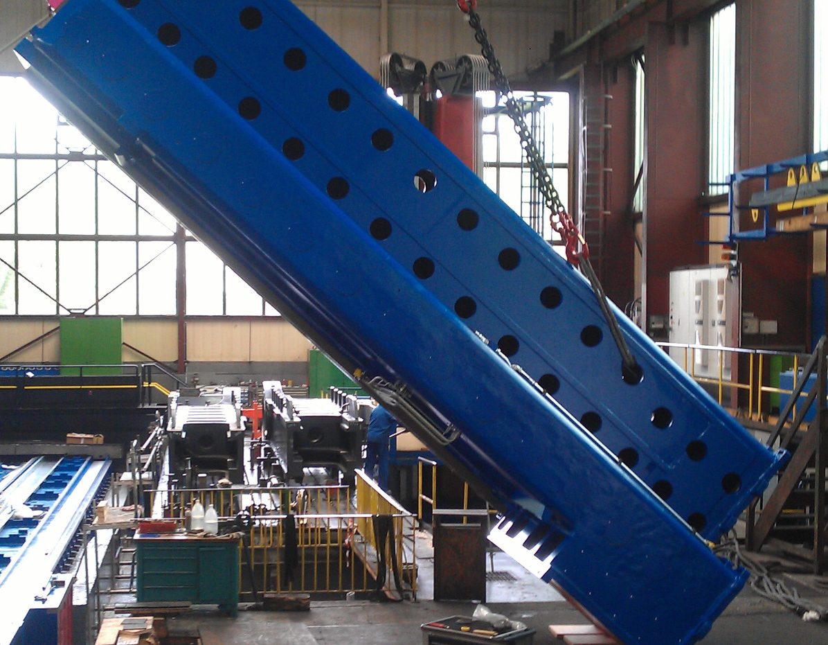 Mitarbeiter bewegen ein großes blaues Maschinenteil mit einem kran in einer Industriehalle